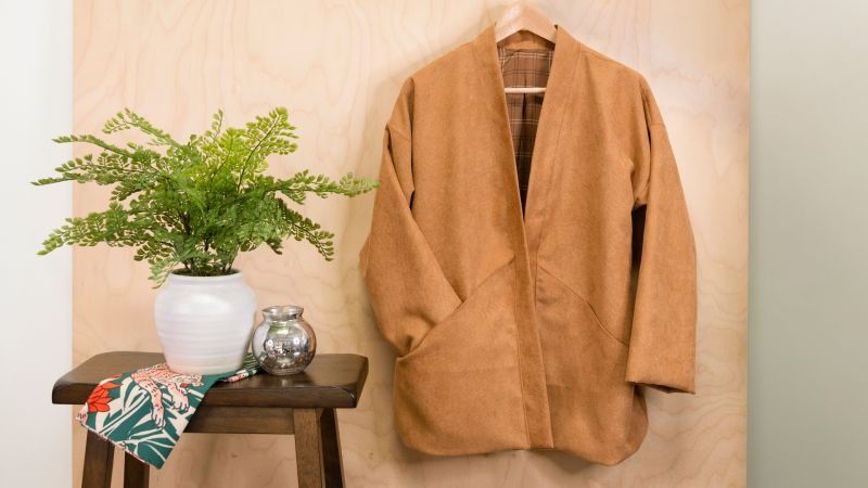 Image of a brown Hanten Coat on a hanger right of a table with a green plant on it.