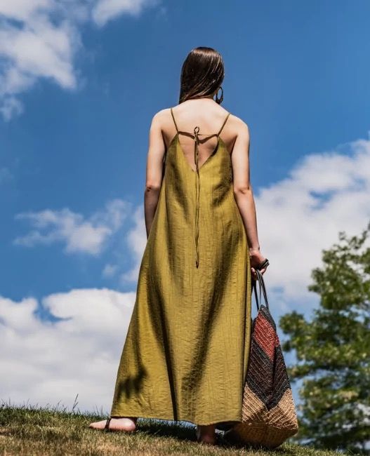 Back view of a woman wearing a maxi-length slip dress which is fitted at the shoulders with spaghetti straps, flaring into a deep vneck at the back, tied with loose back ties in the middle back.
