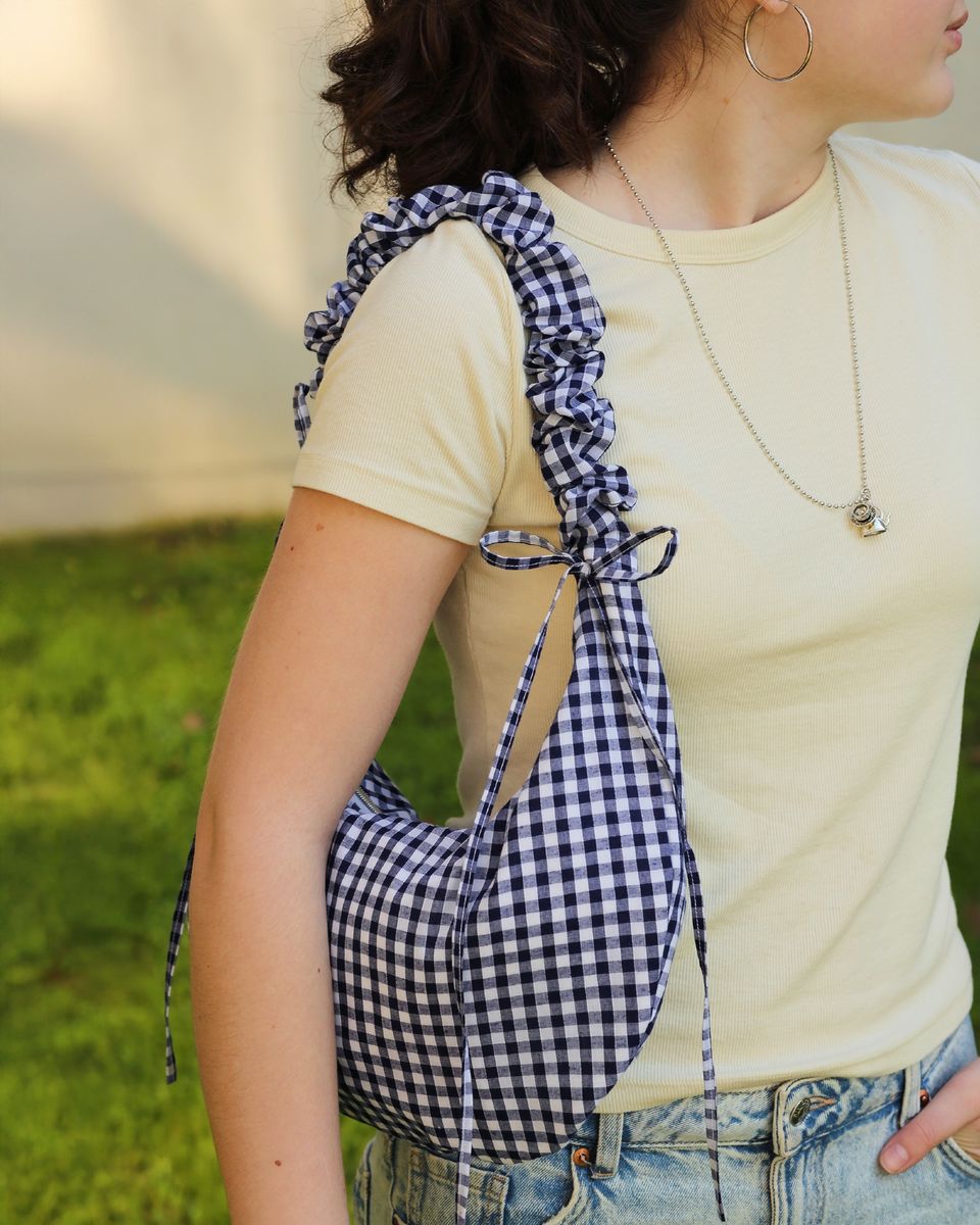 A model is wearing the Drawstring Crescent Bag with the short scrunched up strap on her left shoulder. The ties are tied into bows. The bag is made from a gingham cotton. This bag can be made with the PDF sewing pattern and video tutorial.