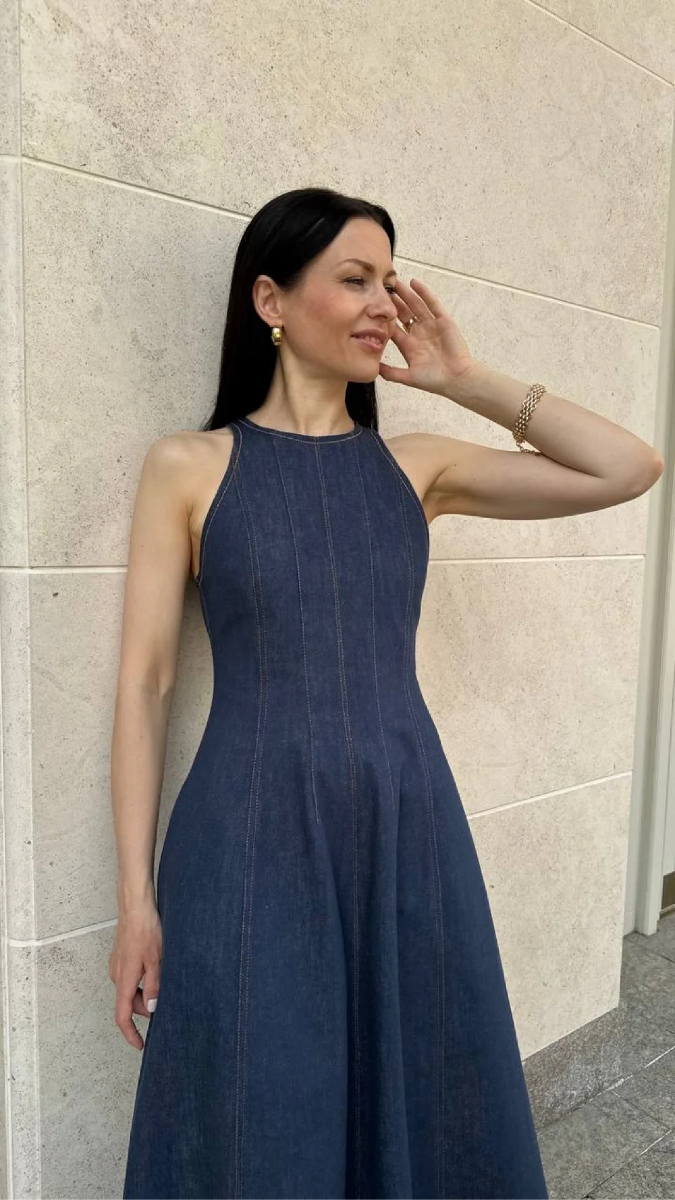 A 3/4 photo showcasing a dark blue sleeveless denim dress on a woman with dark hair and light skin. The dress features princess seams, a high neck, and visible topstitching. 