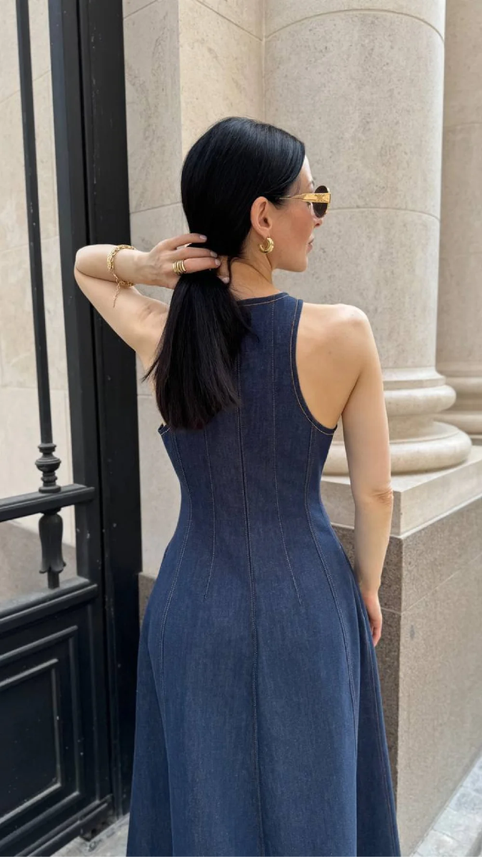 Image of woman with dark hair and light skin facing away from the camera. She is lifting her long hair away with her left arm to show off the invisible zipper sewn onto a dark blue denim dress. 