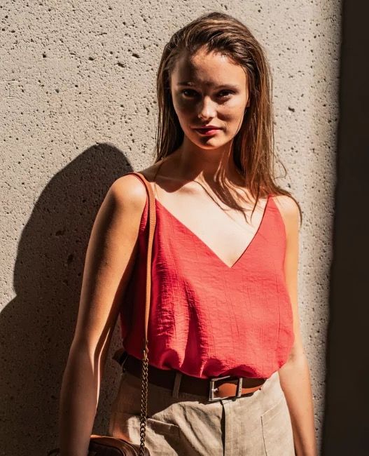 Woman wearing a sleeveless top in a textured red fabric which is fitted at the shoulders with short spaghetti straps, extending into a deep vneck at the front. The top is tucked into trousers.