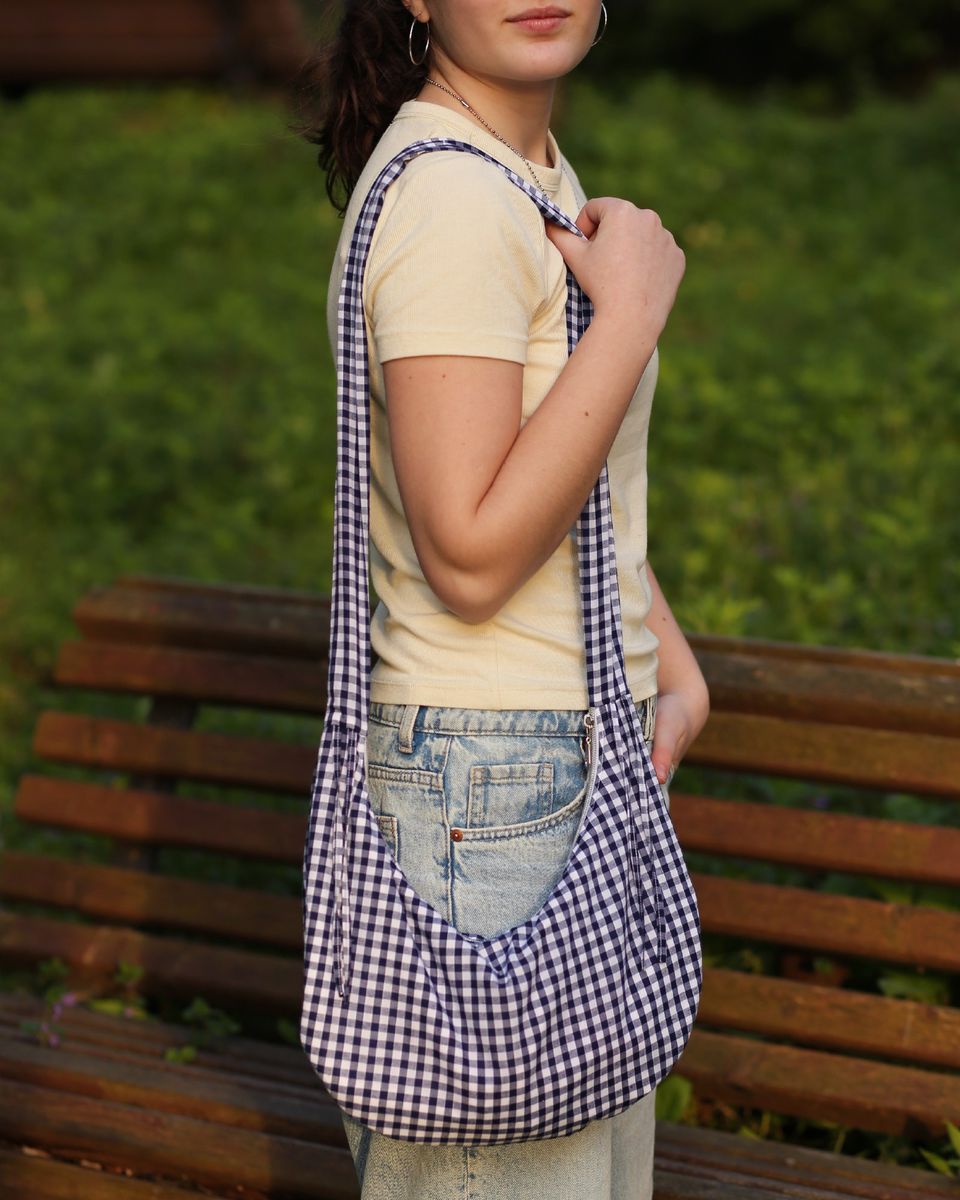 A model is wearing the Drawstring Crescent Bag with the long strap on one shoulder, like a messenger bag. The ties are tied into bows. The bag is made from a gingham cotton. This bag can be made with the PDF sewing pattern and video tutorial.