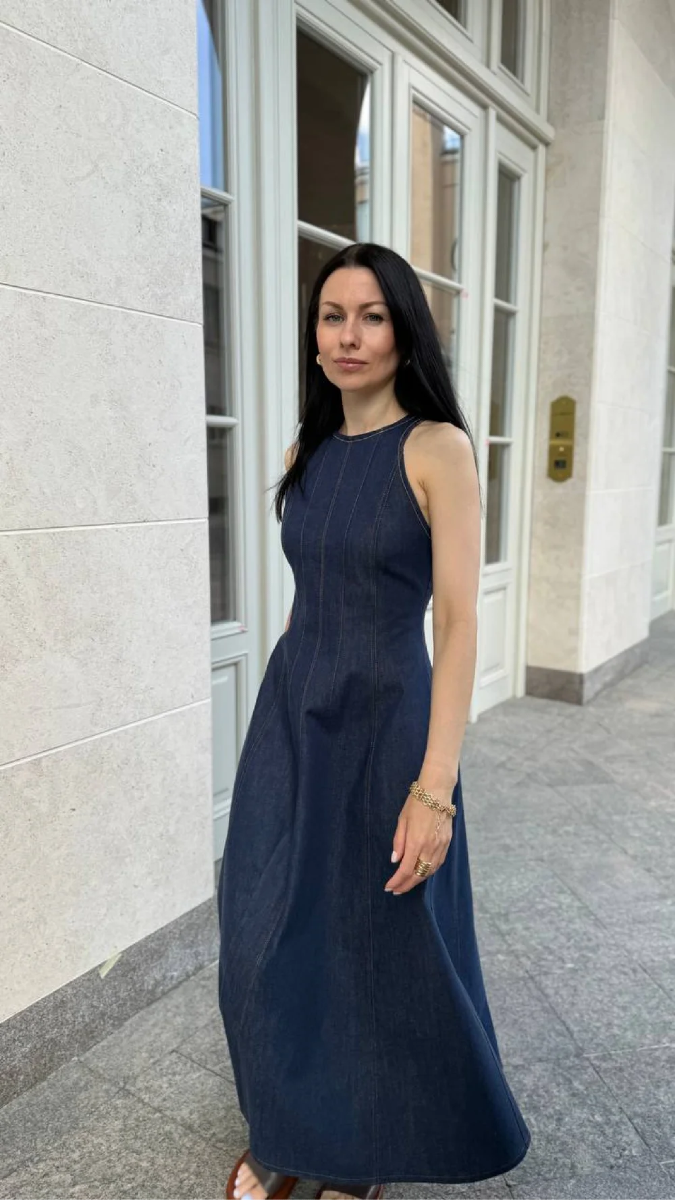 Image showing woman with dark hair and light skin walking past a camera. She is wearing a blue denim dress with visible top stitching and princess seams. 