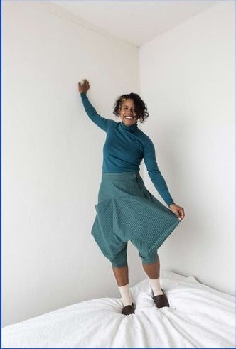 Person in teal Leija trousers standing in a white area.  They are holding one side of the trousers to show the interesting shape.